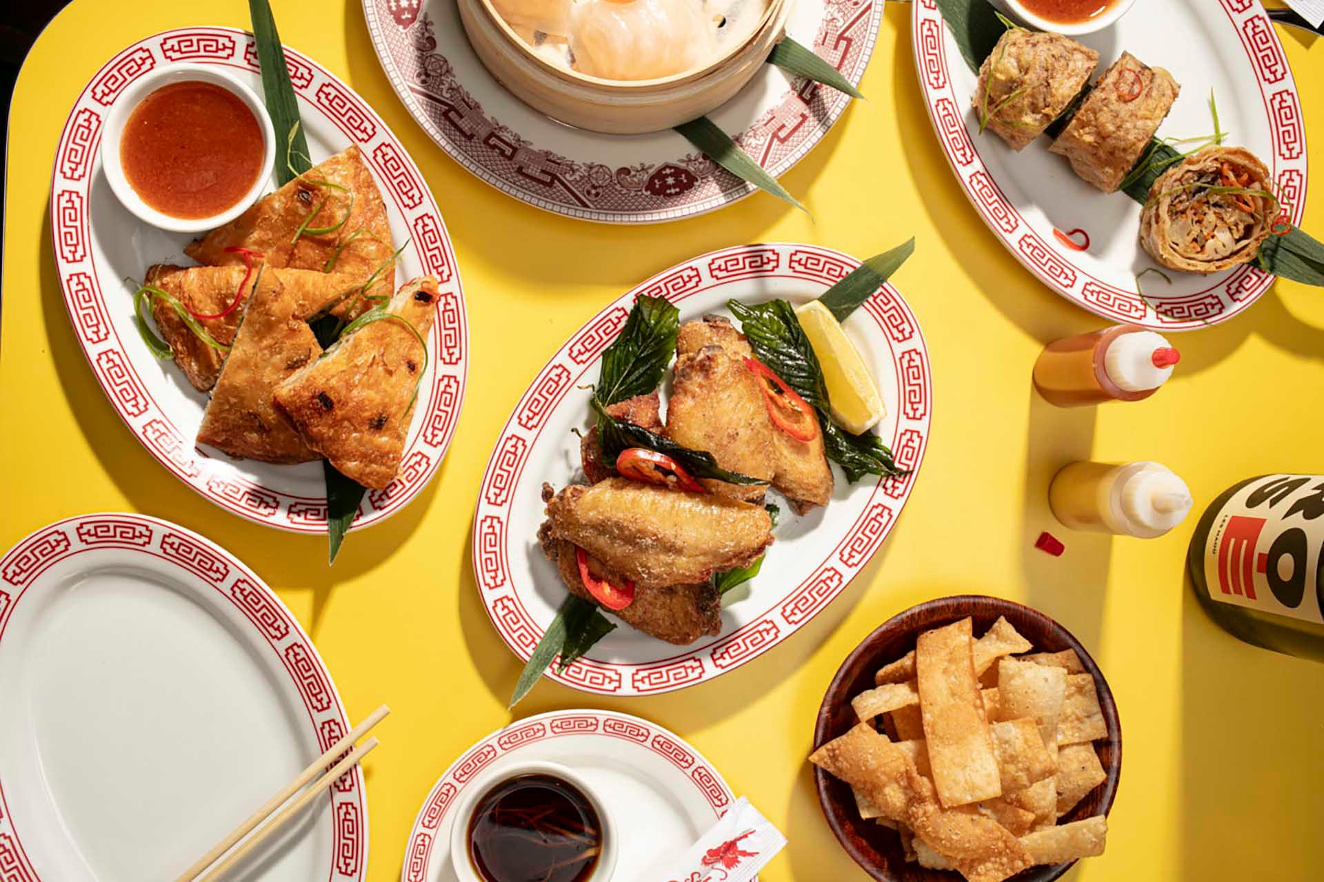 A vibrant table with a variety of Asian dishes on decorative plates, including dim sum, spring rolls, and fried snacks. Condiment bottles and dipping sauces accompany the dishes on a yellow background, creating a colorful dining scene.