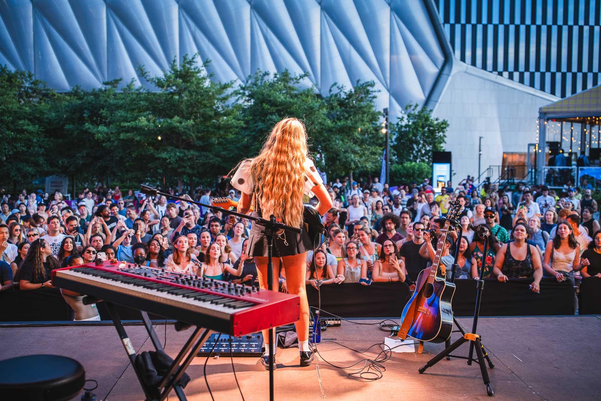 A musician with long hair performs on an outdoor stage, facing a large crowd. Musical instruments, including guitars, are nearby. Trees and a modern building are visible in the background.