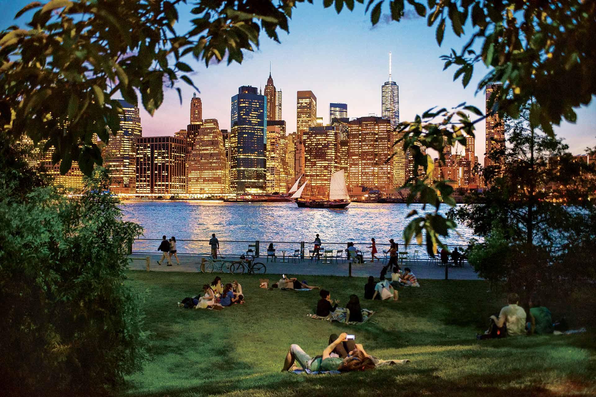 People relax on a grassy park overlooking a river, with a sailboat and the illuminated New York City skyline in the background at dusk. Trees frame the view in the foreground.