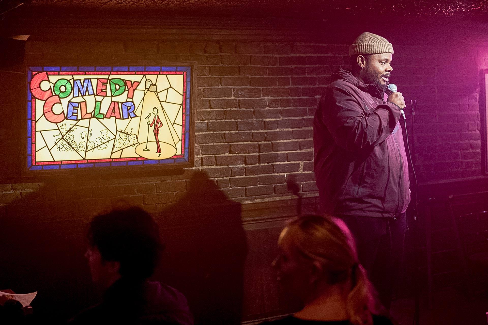 A comedian performs onstage with a microphone at the Comedy Cellar, standing beside a colorful stained-glass sign, while audience members sit and watch in a dimly lit, brick-walled room.