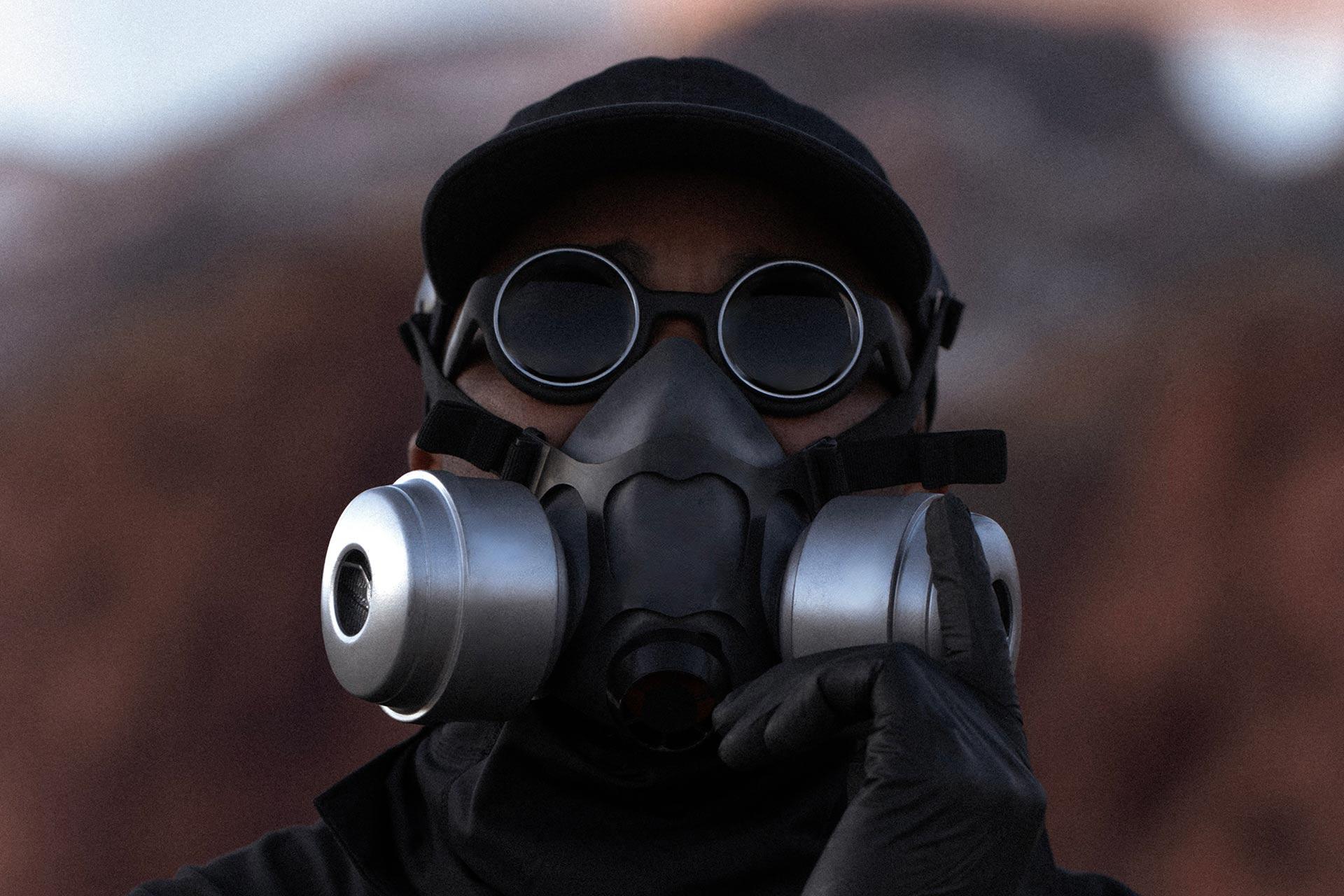 A person wearing dark round goggles, a black cap, and a black gas mask with large silver filters, dressed in black clothing and gloves, stands outdoors with a blurred rocky background.
