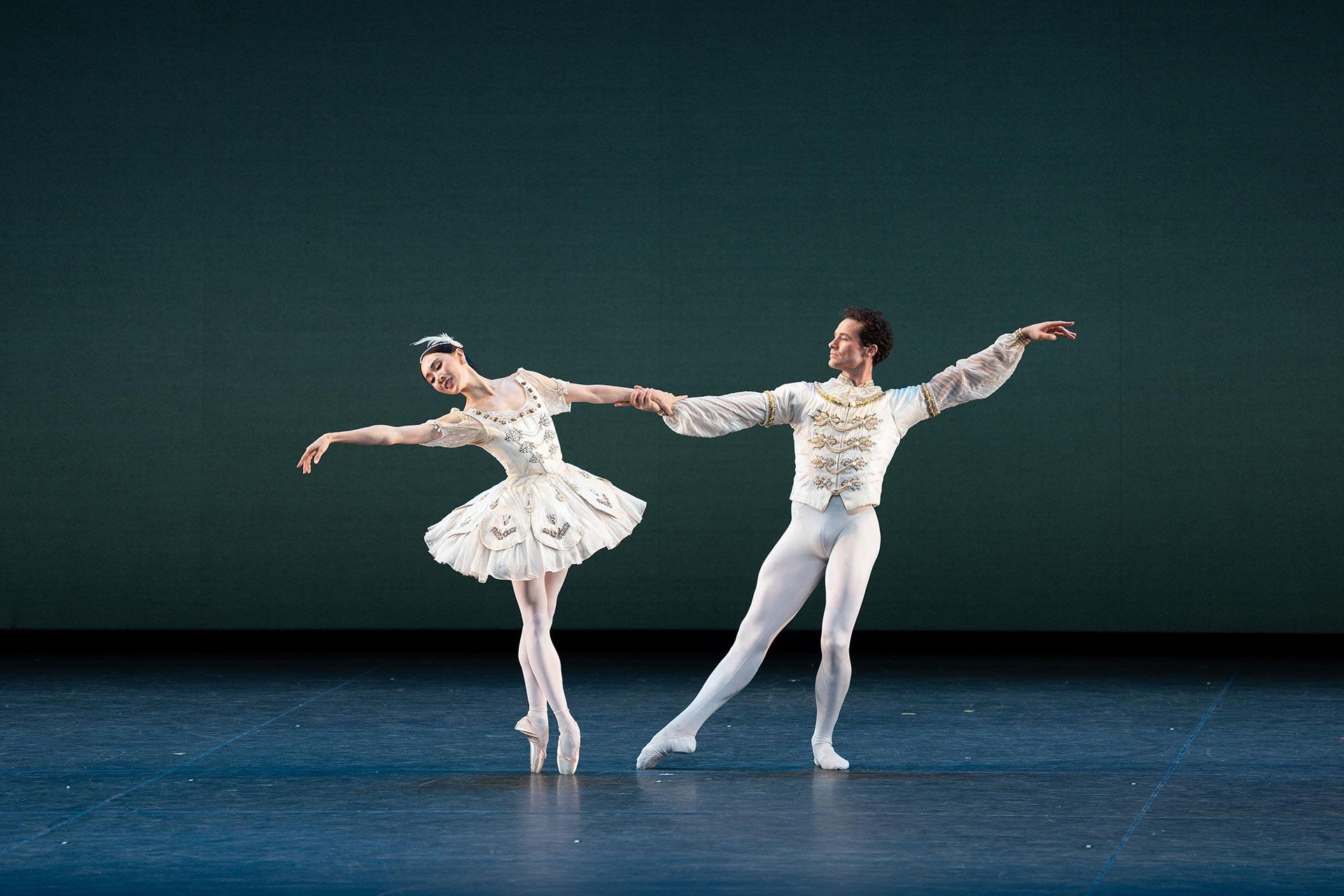 Two ballet dancers in white costumes perform on stage. The female dancer balances en pointe with one arm extended, while the male dancer holds her hand, both poised gracefully against a plain backdrop.