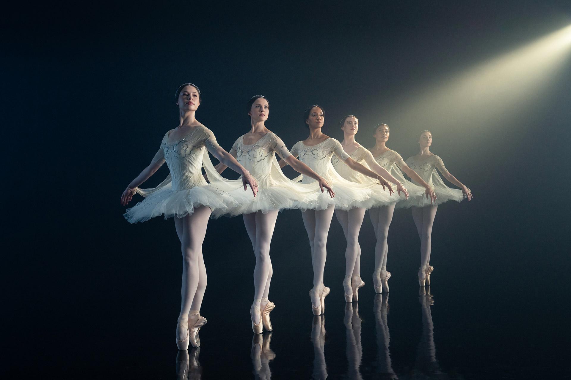 Five ballerinas in white tutus stand in a line on pointe, arms outstretched and slightly curved, performing on a dark stage with a spotlight shining from the upper right.