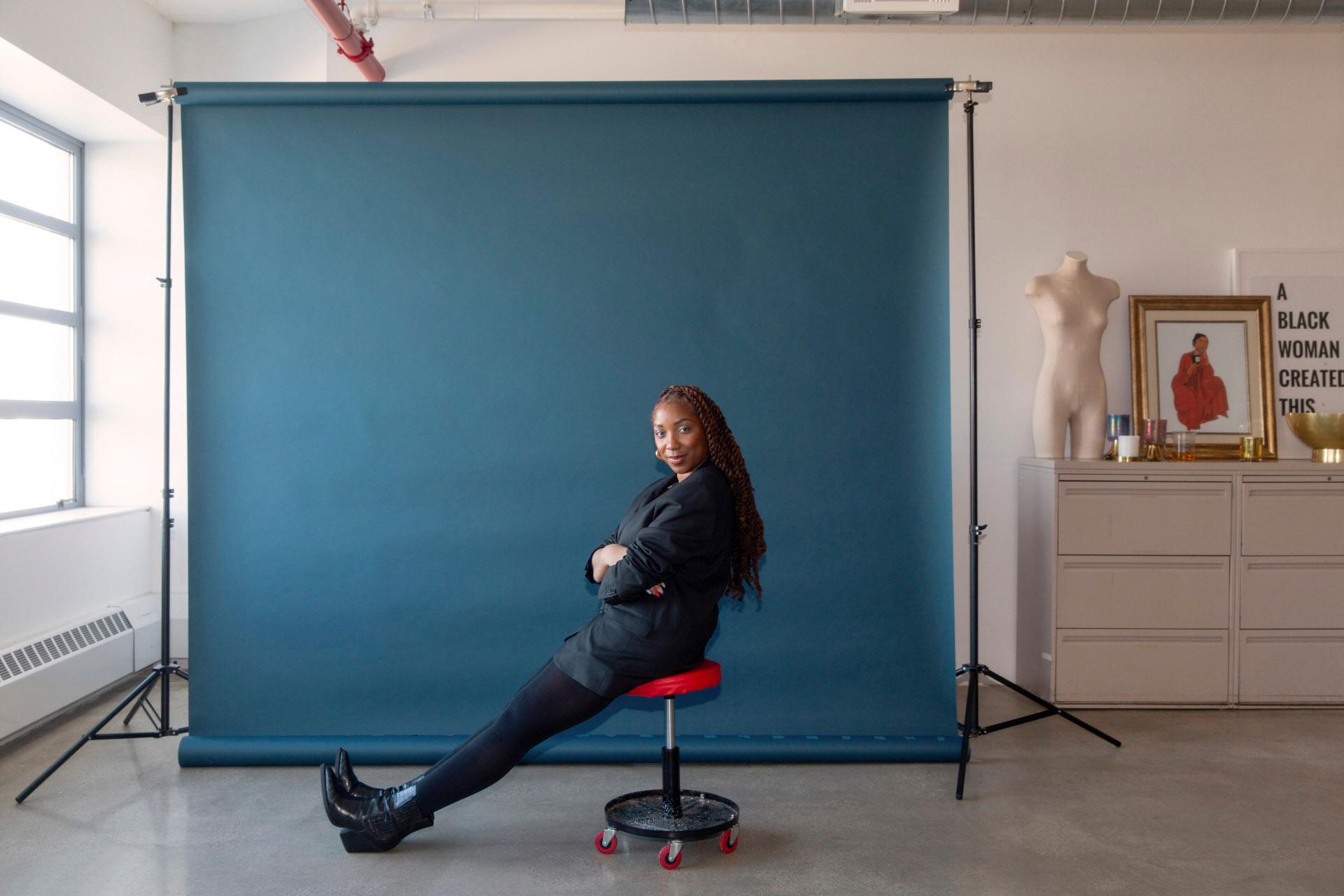 A person with long, braided hair sits on a red stool, wearing a black outfit. They are in front of a dark blue backdrop in a bright room. Nearby is a dresser with a torso mannequin and a framed text that reads, "A BLACK WOMAN CREATED THIS.