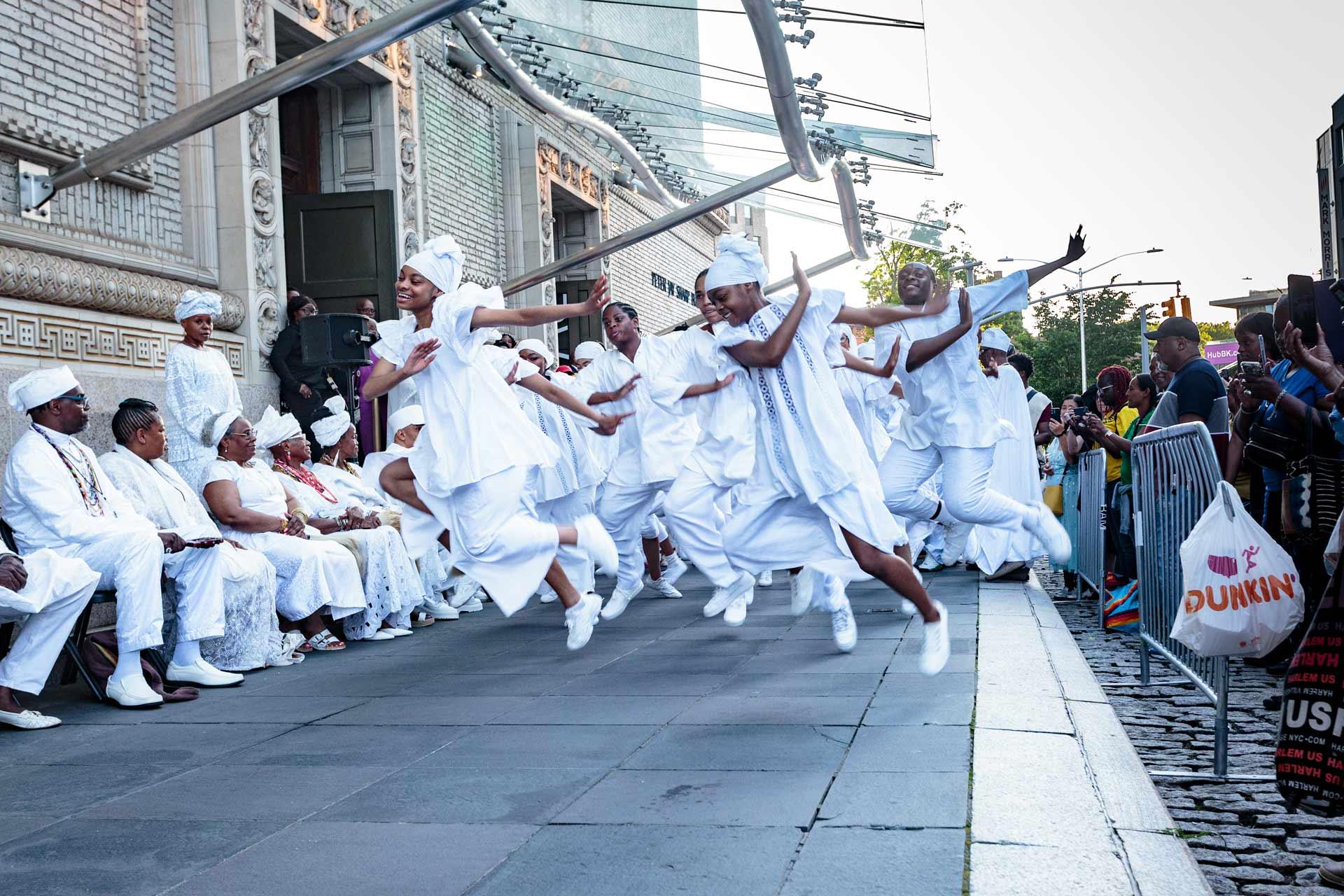Dancers in white outfits perform energetically in an outdoor urban setting. A group of seated people in white watch the performance, while a crowd on the right observes, separated by barriers. The event takes place under a clear sky.