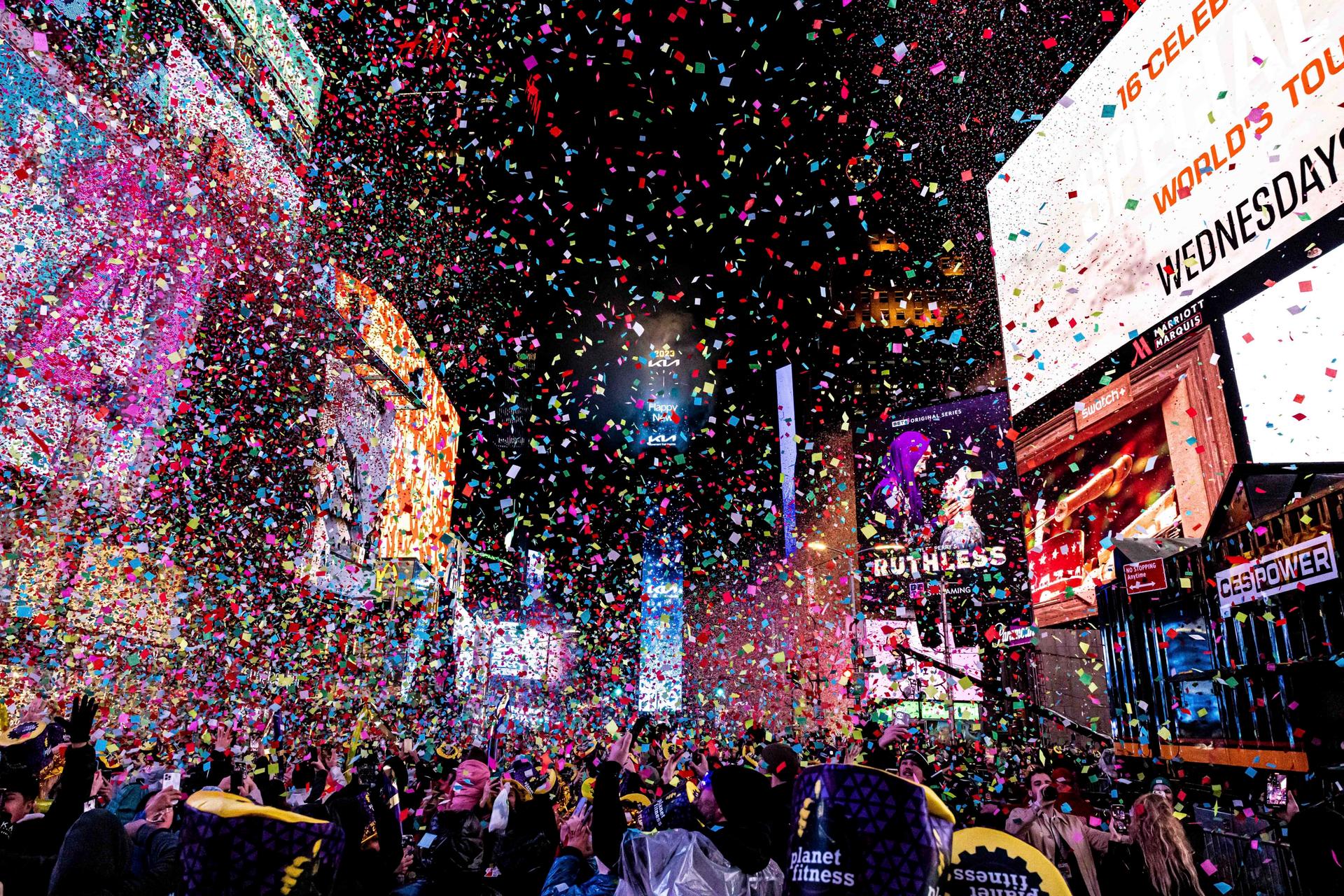 A large crowd celebrates in Times Square at night as colorful confetti fills the air, surrounded by bright electronic billboards and city lights.