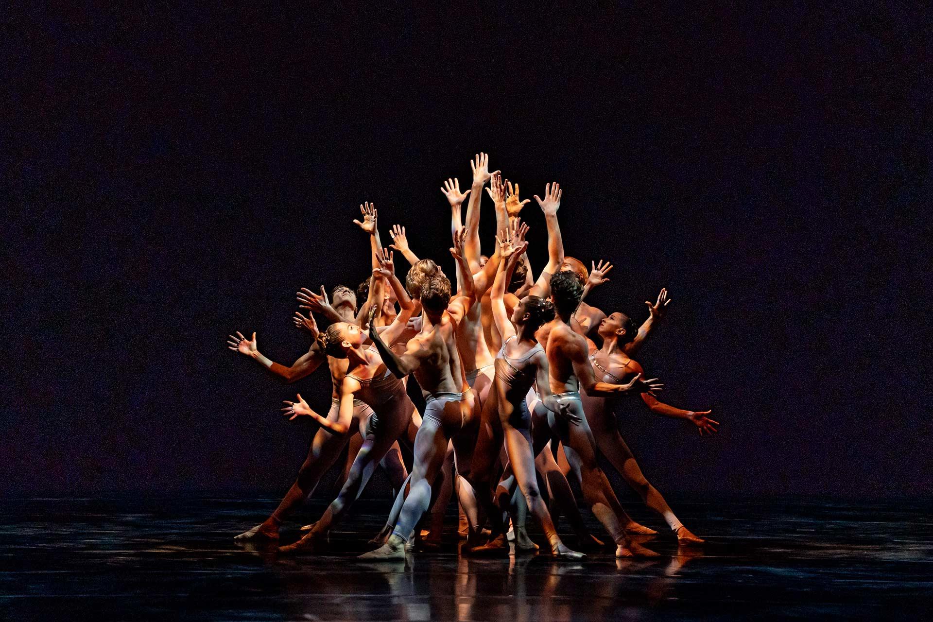 A group of dancers on stage form a tight circle, reaching their arms upward and outward. They wear skin-toned costumes and are dramatically lit against a dark background, creating a striking visual effect.