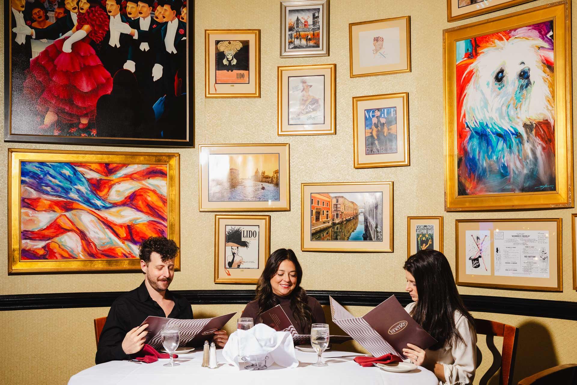 Three people sitting at a round table in a restaurant, looking at menus. The walls are decorated with various framed paintings and photographs, including a dog and abstract art. The table is set with glasses and a white tablecloth.