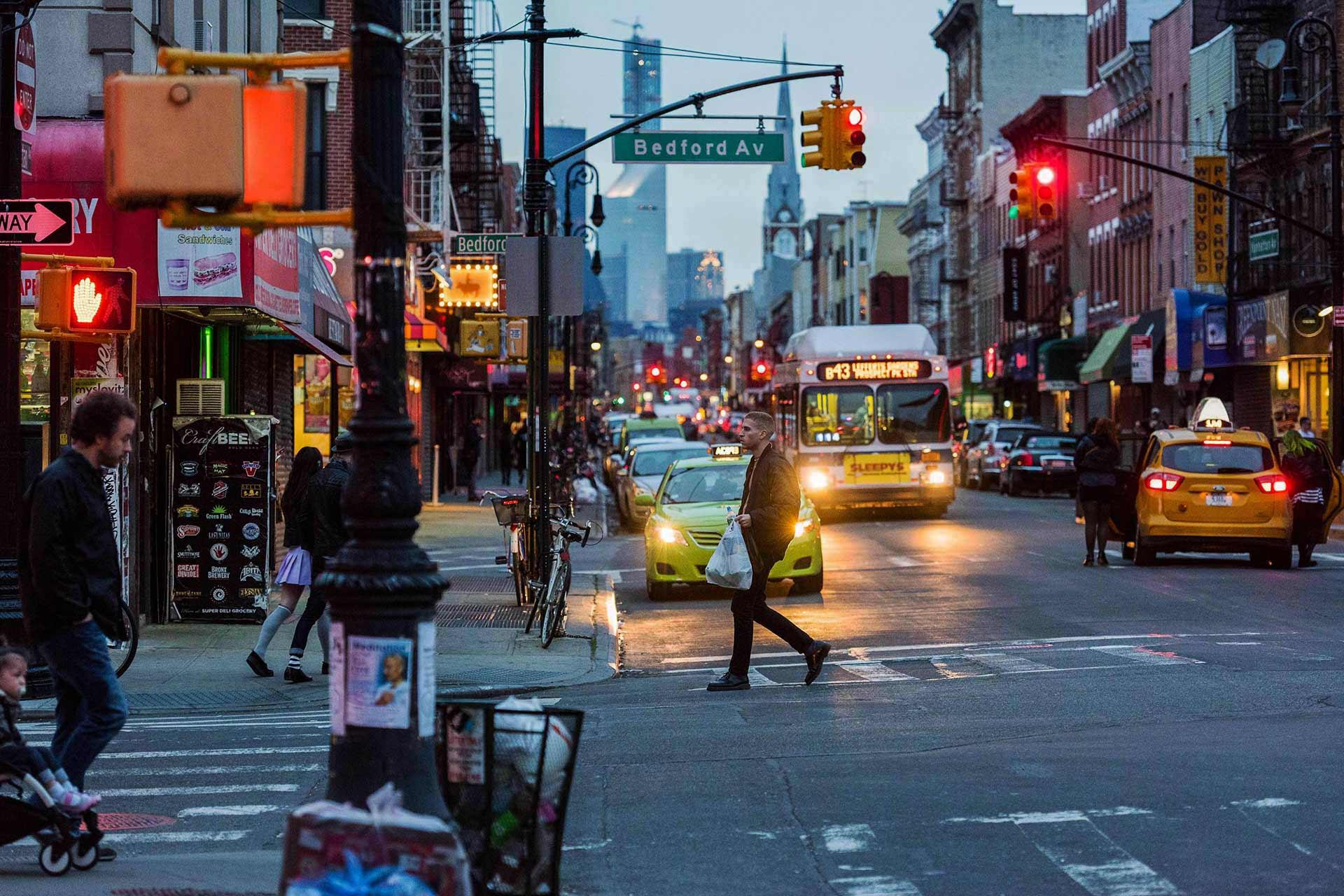 People cross a busy city street at dusk, with cars, a bus, yellow taxis, and buildings lining both sides. Traffic and street lights are illuminated, and a street sign reads "Bedford Av.