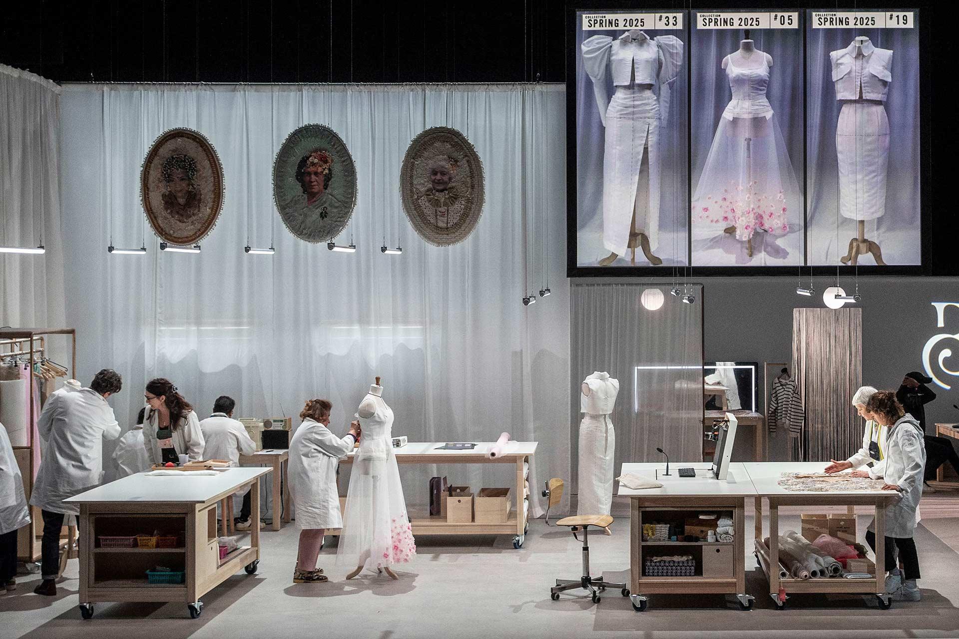 Fashion designers in white coats work on garments at large tables in a studio. Mannequins, sketches, fabrics, and finished dresses are on display, with photos of spring 2023 designs hanging above.