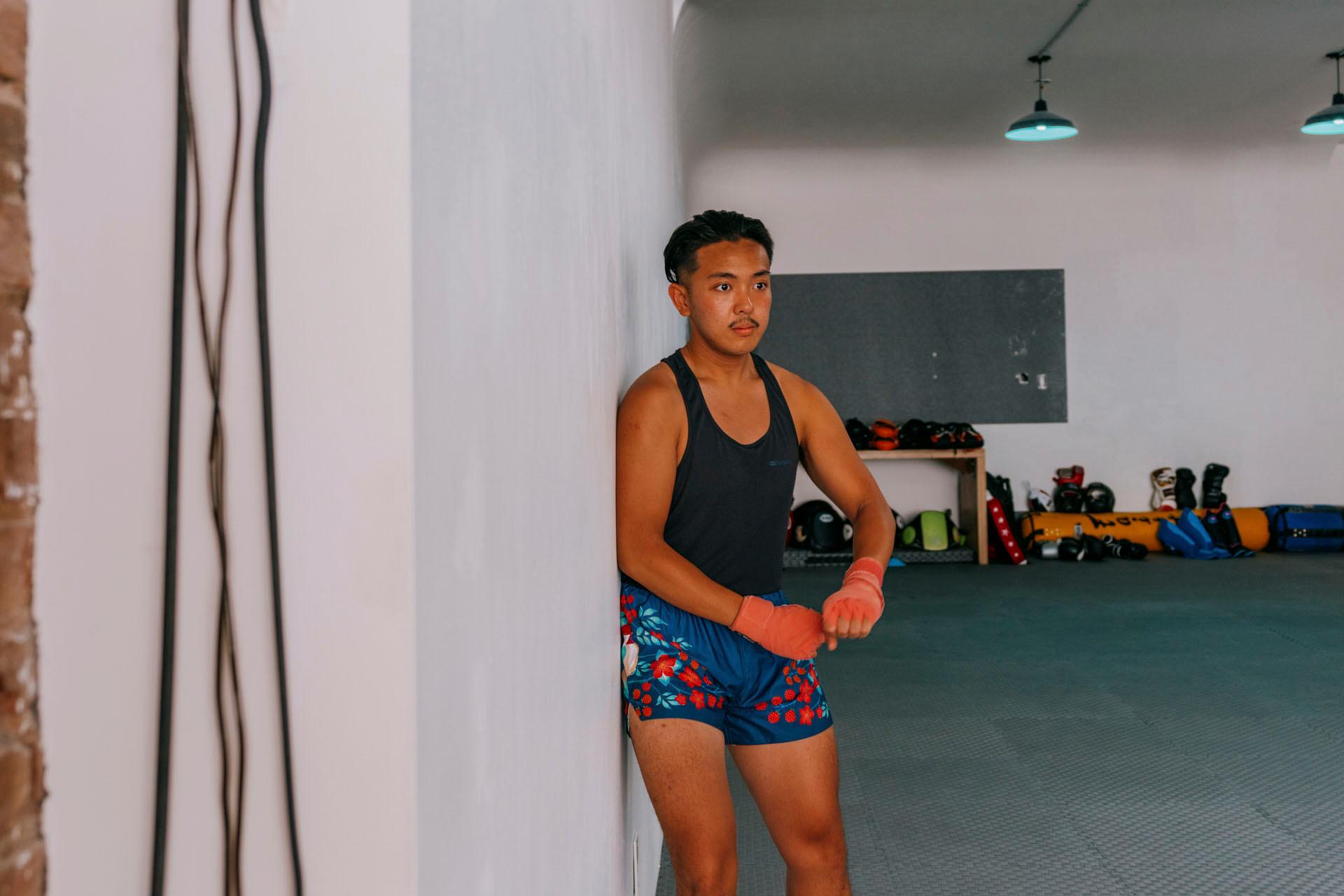 A person with long hair is leaning against a white wall in a gym, wearing a dark tank top and patterned shorts. Their hands are wrapped, and they appear focused. Gym equipment is visible in the background.