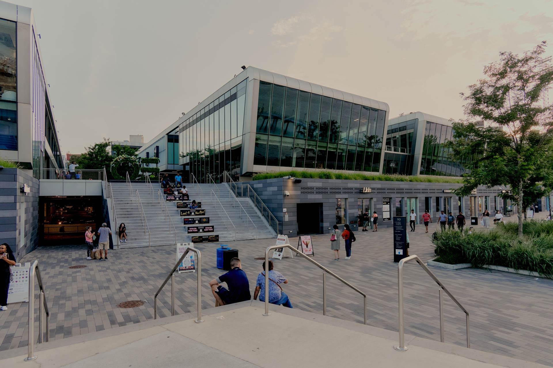 Modern university building with large glass windows, outdoor steps, people walking and sitting, and greenery around the entrance on a paved plaza. The sky is cloudy and the scene is lively with activity.