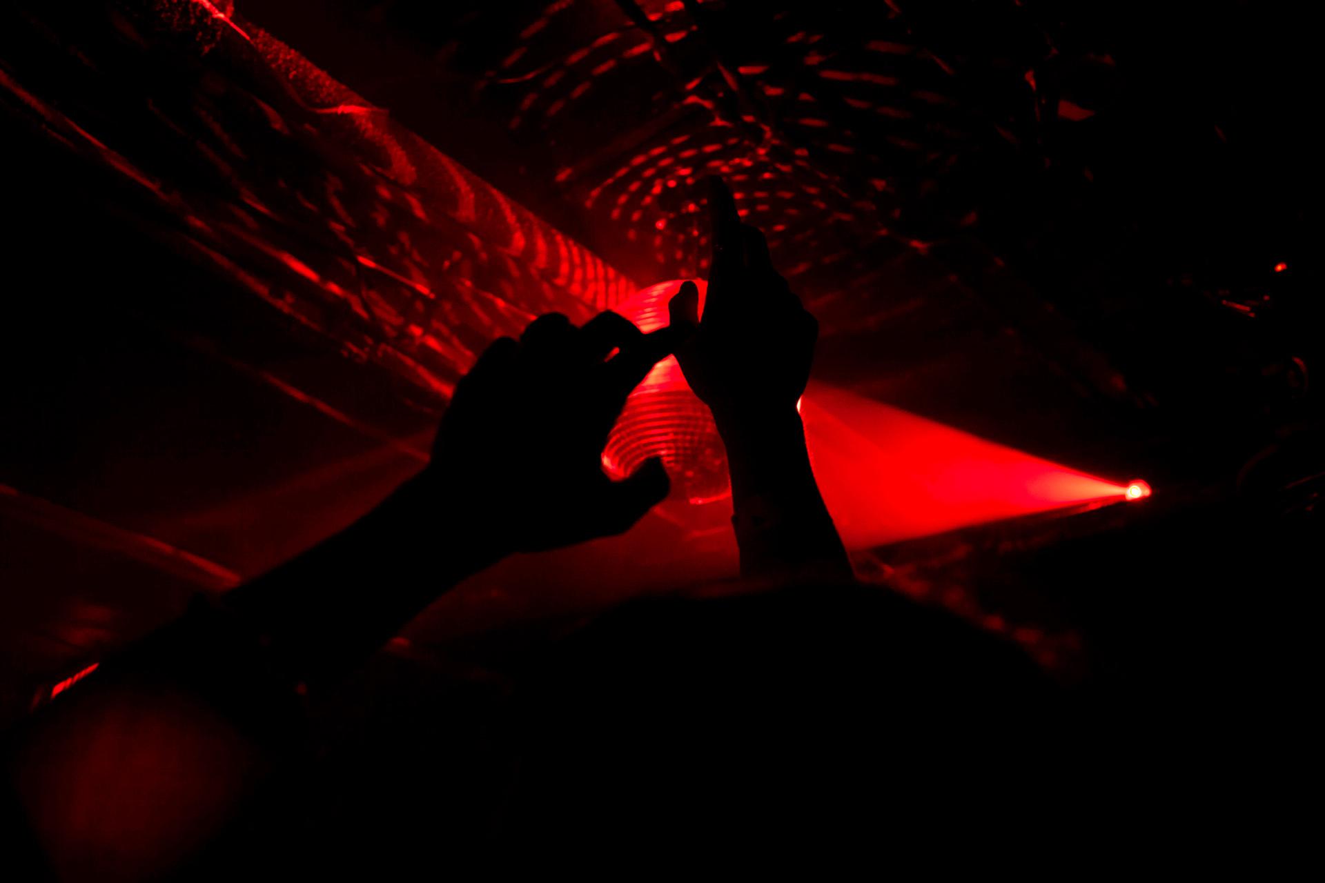 Silhouetted hands are raised against a backdrop of red lights and laser beams at a concert or club. The dark atmosphere creates a dramatic contrast with the vibrant red glow.