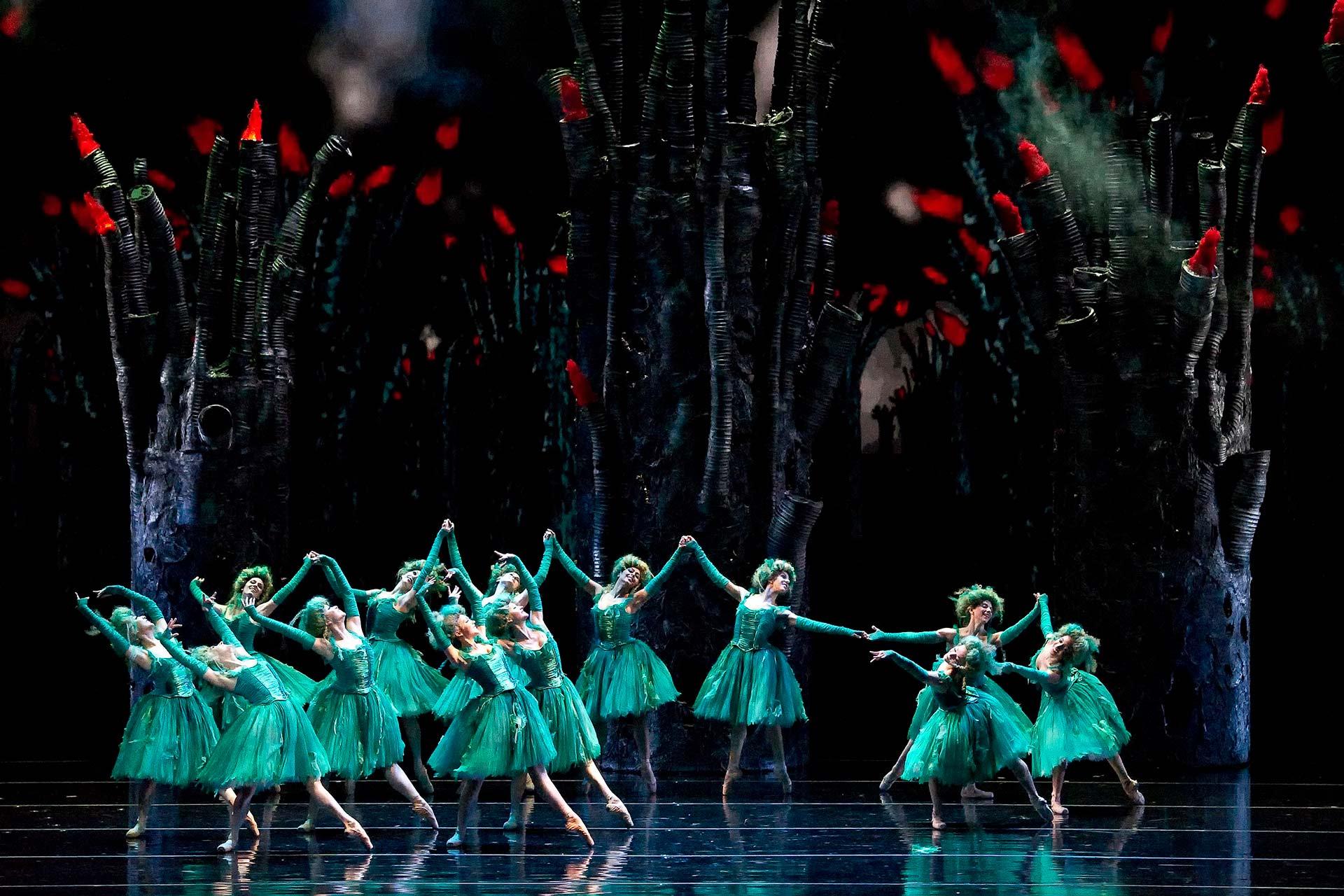 Ballet dancers in matching green costumes perform on stage, holding hands in a circle. The background features dark, towering abstract trees with red-tipped branches, creating a mystical, enchanted forest scene.