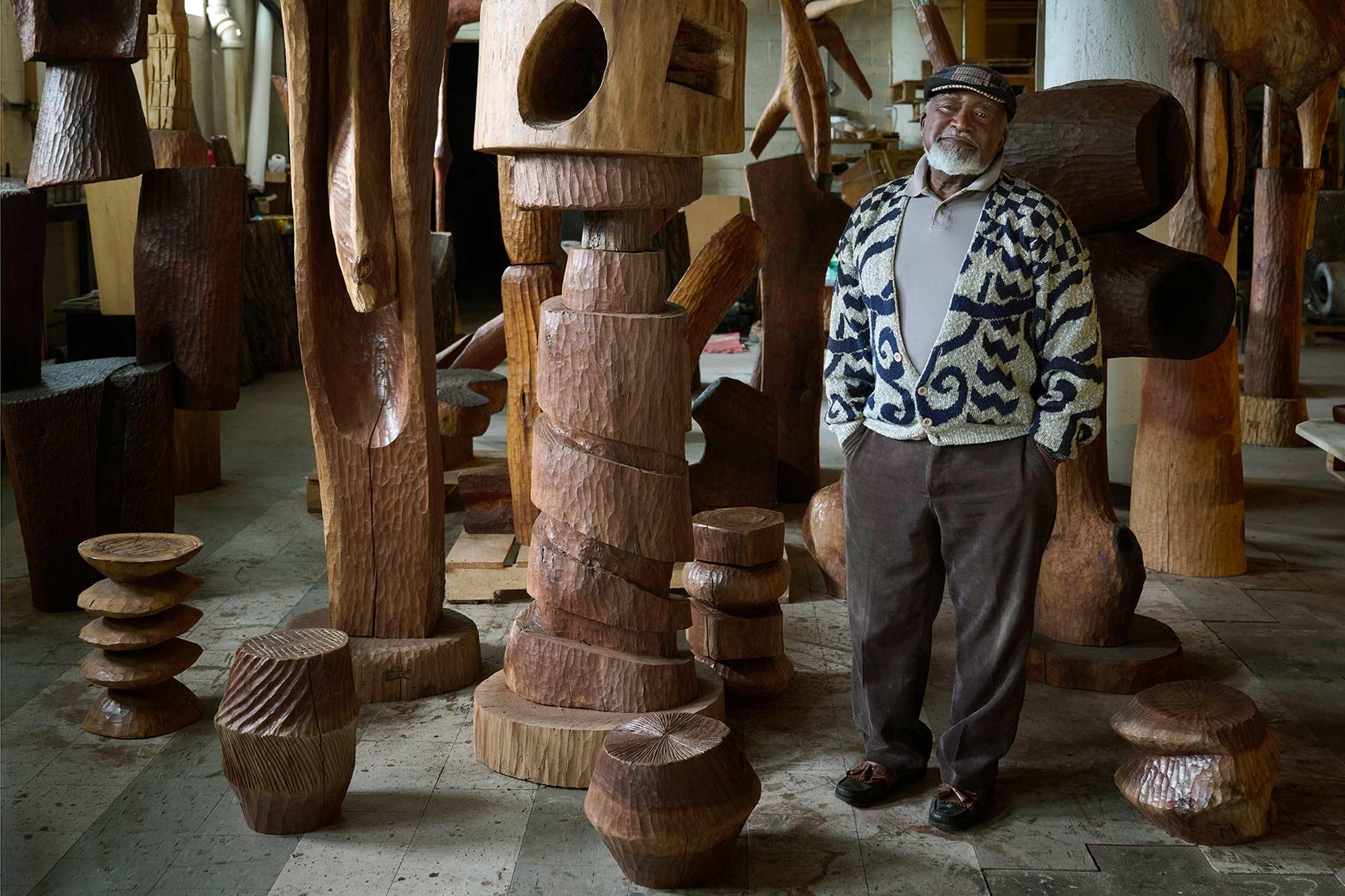 An older man in a patterned sweater and cap stands in a workshop surrounded by large, abstract wooden sculptures and carved wooden stools. The space is filled with natural light and unfinished wooden artworks.