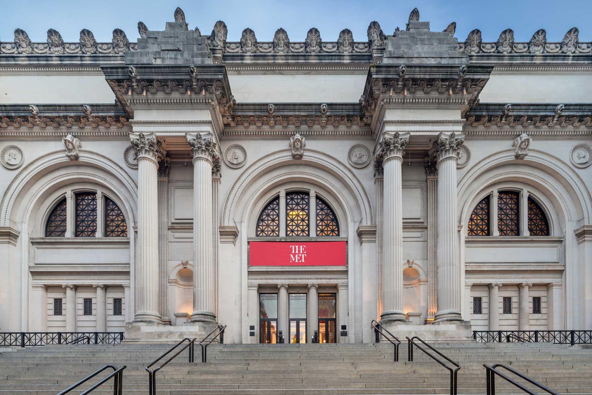 Facade of a grand museum with neoclassical architecture featuring large columns, arched windows, and ornate detailing. A red banner with "The Met" hangs above the entrance. Stone steps lead up to the entrance.