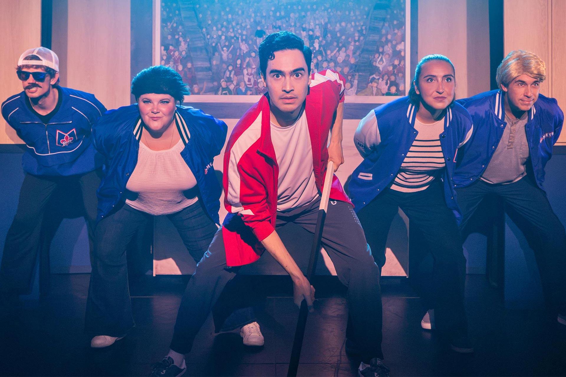 Five people in sports jackets pose dramatically on stage, with one person in the center holding a hockey stick. They stand close together with a stadium crowd shown on a screen behind them, under blue and pink lighting.