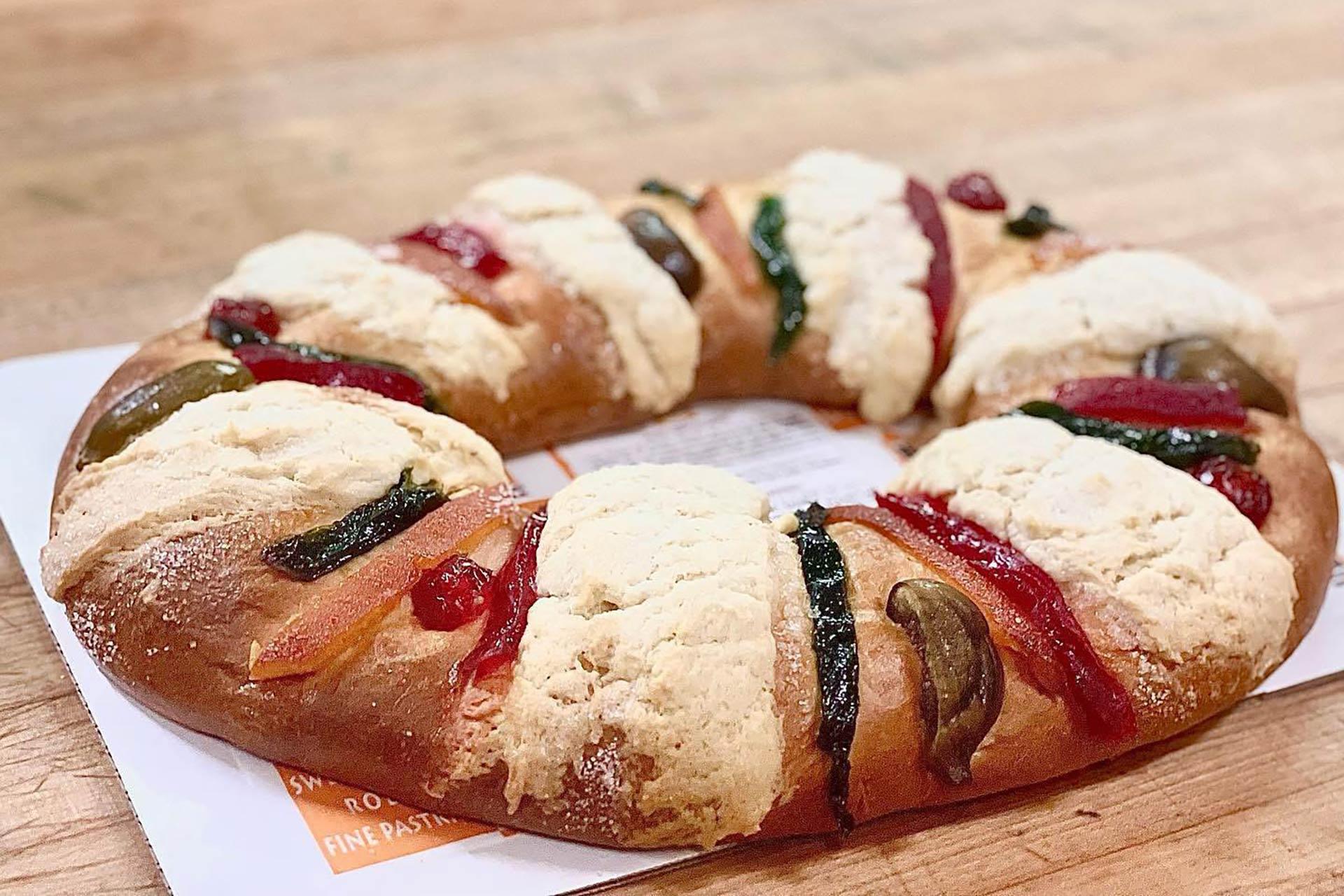A round, ring-shaped sweet bread decorated with colorful candied fruits and sugar topping, placed on a wooden surface.