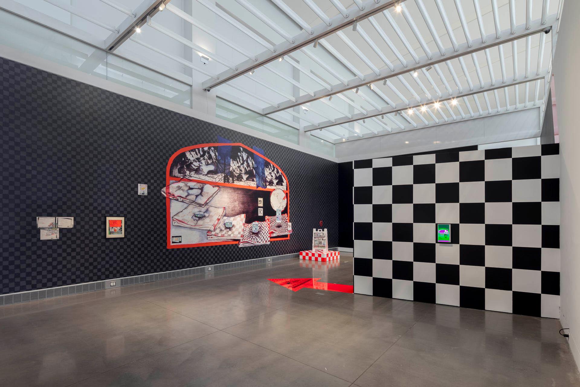 A modern art gallery features a large checkered black-and-white structure, a mural depicting a diner scene on a patterned wall, small framed artworks, and a red striped installation on a shiny concrete floor.